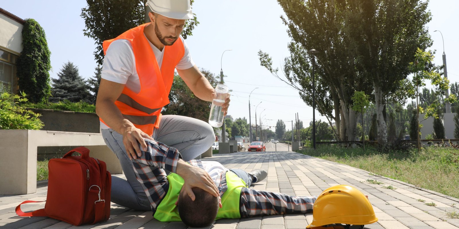 Construction Worker Assisting Colleague with Heat Illness in California Construction worker in an orange vest giving water to an injured colleague sitting on the pavement, illustrating workplace heat illness and emergency response in California.