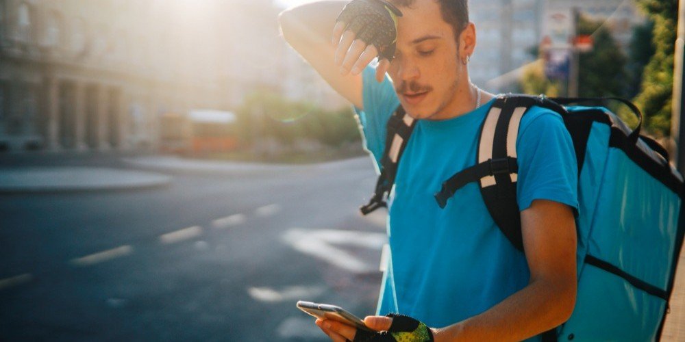 Delivery Worker Experiencing Heat Exhaustion Outdoors in California Young delivery worker wiping sweat from his forehead while checking his phone on a hot sunny day, representing heat exhaustion risks in California workplaces.