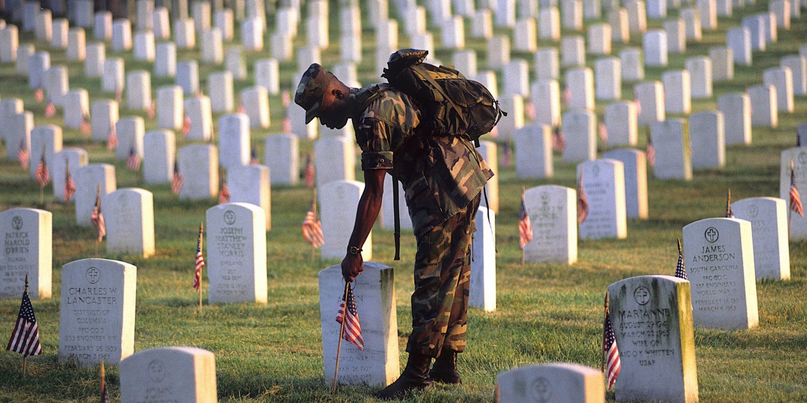 Memorial Day Tribute at Military Cemetery A soldier in uniform respectfully places a small American flag on a headstone in a military cemetery filled with white grave markers and flags, symbolizing remembrance of fallen heroes on Memorial Day.