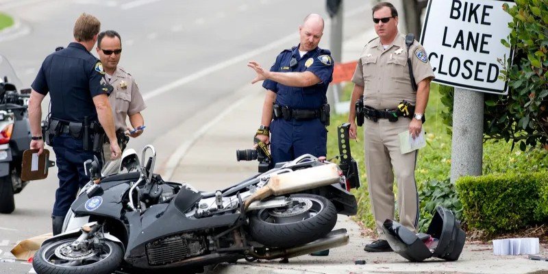 Group of police officers standing around a crashed motorcycle on a city street next to a 'Bike Lane Closed' sign, documenting the accident scene.