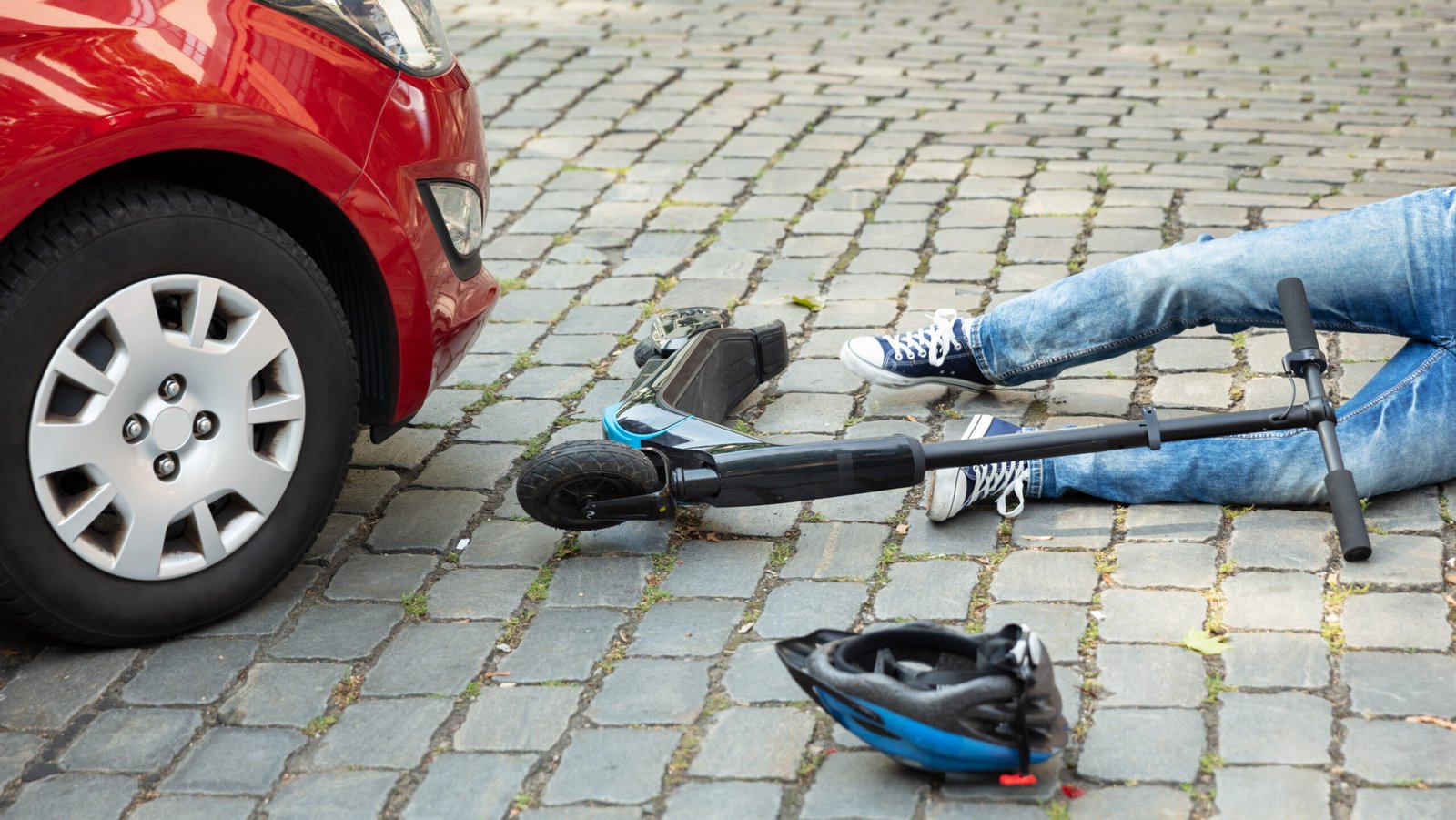 A person lying on the ground after a collision between a red car and an e-scooter in a city street, with a helmet and scooter on the ground.