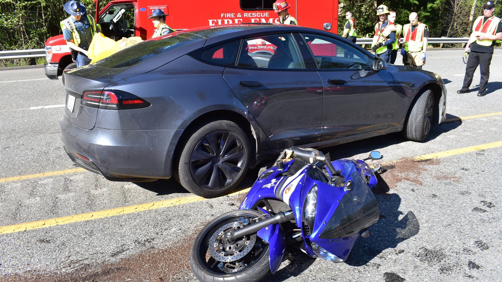 A Tesla car involved in a crash with a motorcycle, with emergency responders on-site, highlighting potential liability issues in self-driving car accidents.