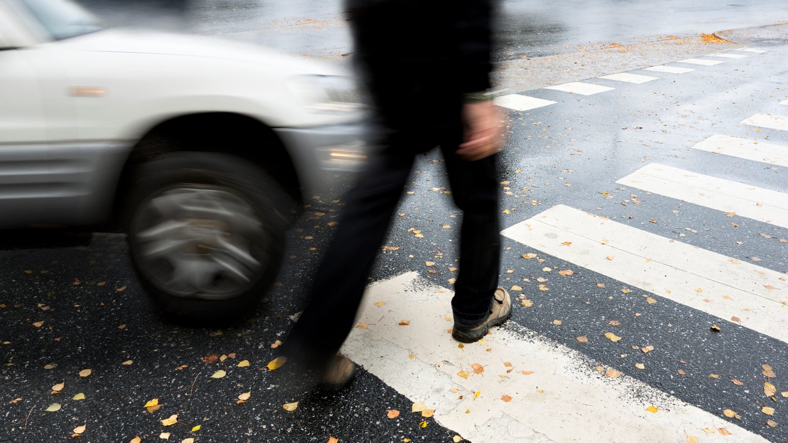A pedestrian walking across a wet crosswalk as a car approaches closely, showing the potential risk factors involved in a pedestrian accident.