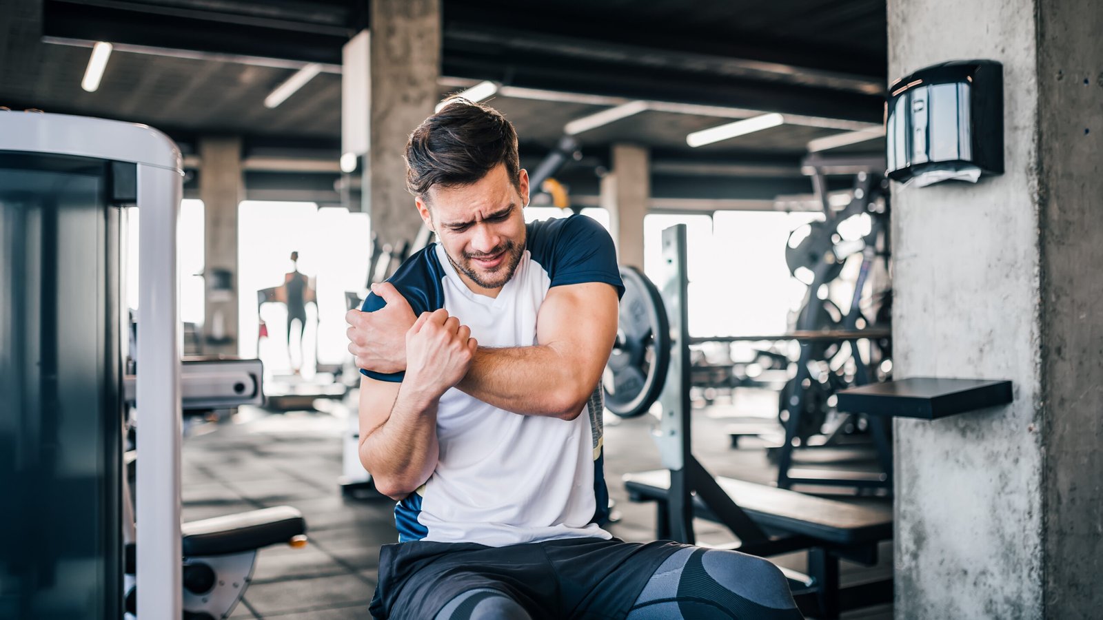 Man sitting in a fitness center holding his arm in pain after being injured at a gym, highlighting common workout-related injuries.