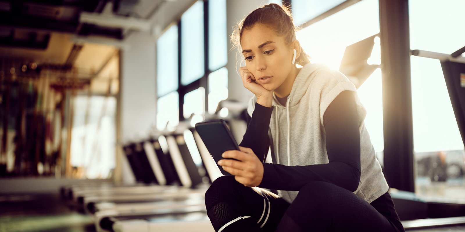 Concerned woman sitting in a gym after a workout injury, looking at her phone to research legal options for being injured at a gym.