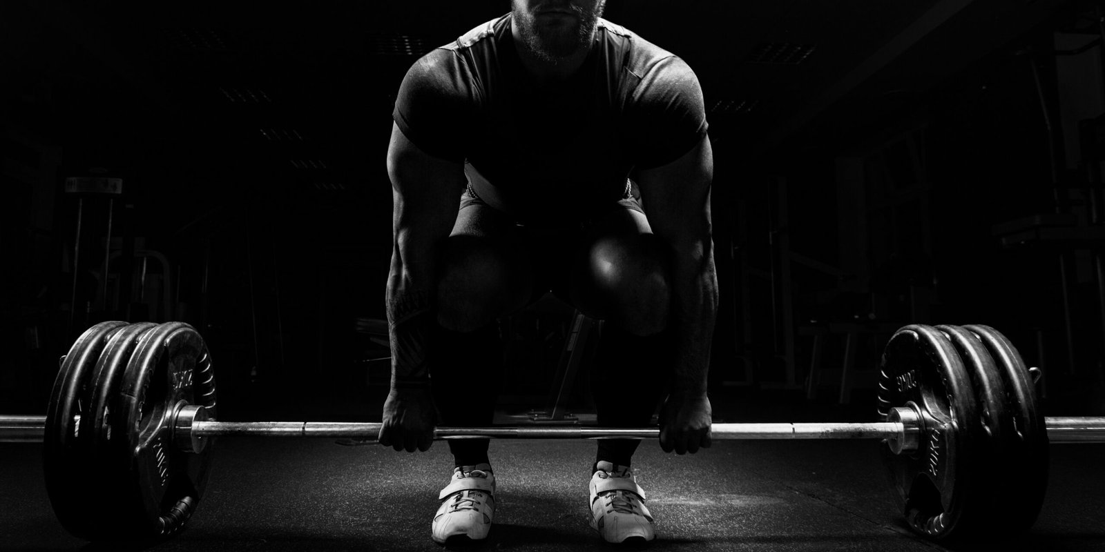 Black and white image of a muscular man preparing to lift a heavy barbell in a dimly lit gym, representing the discipline and strength powerlifting brings to personal injury law.
