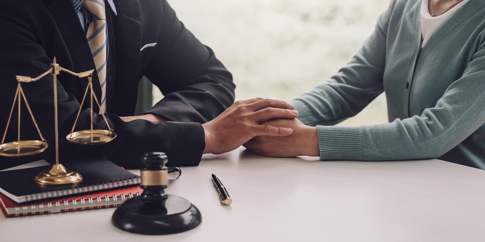 A lawyer in a suit gently holds a client's hand across a desk with legal documents and a justice scale, representing The Powerlifting Lawyer's compassionate approach to personal injury cases.